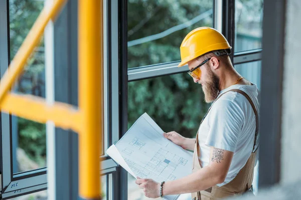 Vista laterale del costruttore in hardhat e googles protettivi guardando cianografia in cantiere — Foto stock