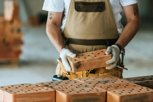 Обрезанное изображение строителя в защитных перчатках с кирпичом на строительной площадке — стоковое фото