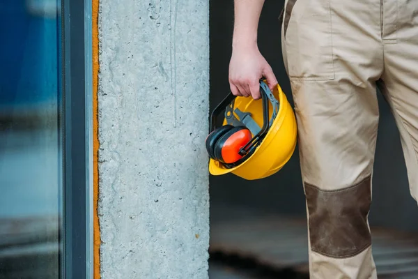 Cropped image of builder in uniform holding construction headphones and protective helmet — Stock Photo