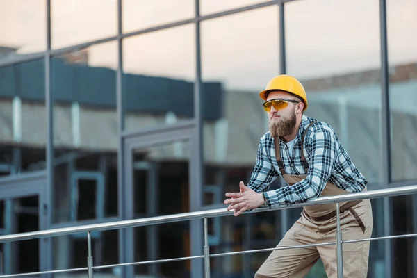 Jeune constructeur dans googles protecteurs et hardhat tenant les mains ensemble près du bâtiment — Photo de stock