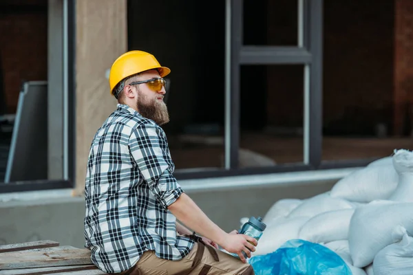 Vista lateral do construtor em óculos de proteção e hardhat descansando com garrafa de água no local de construção — Fotografia de Stock