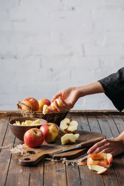 Tiro recortado de mujer con trozos de manzana en la mano y manzanas frescas en la tabla de cortar en la mesa de madera - foto de stock