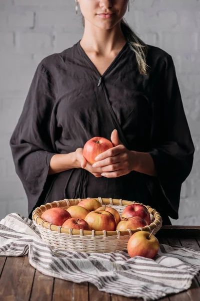 Teilansicht einer Frau mit Apfel und Korb voller frischer Äpfel auf Holztischplatte mit Leinen — Stockfoto