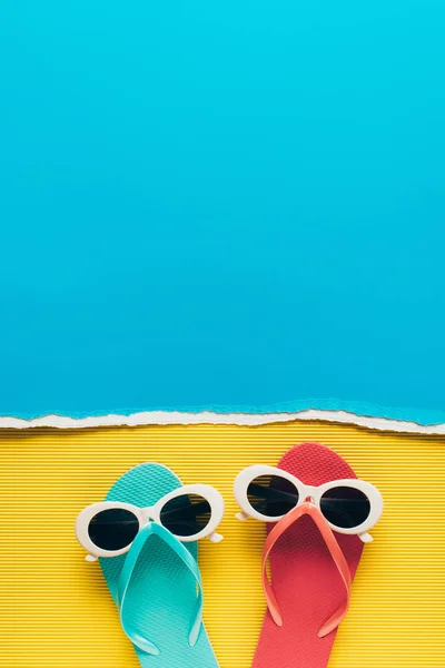 Draufsicht auf blaue und rosa Flip Flops mit Sonnenbrille auf gelb-blauem Papierhintergrund — Stockfoto