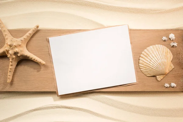 Tendido plano con papel en blanco, estrella de mar y conchas marinas sobre tabla de madera sobre arena - foto de stock