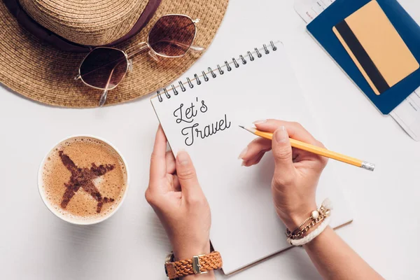Vista parcial de la mujer con cuaderno con permite viajar letras en la mesa con taza de café, sombrero de paja, pasaporte y billete, concepto de vacaciones - foto de stock