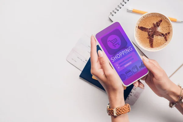 Foto recortada de la mujer con el teléfono inteligente con el logotipo de compras en la pantalla en la mesa con taza de café, billete y pasaporte - foto de stock