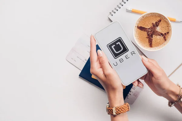 Cropped shot of woman with smartphone with uber logo on screen at tabletop with cup of coffee, ticket and passport — Stock Photo