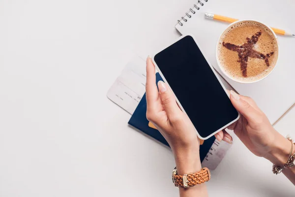 Partial view of woman holding smartphone with blank screen at tabletop with notebook, cup of coffee and passport, traveling concept — Stock Photo