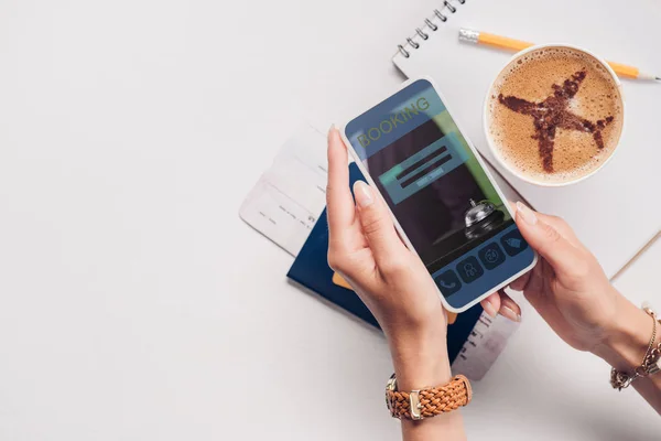 Cropped shot of woman with smartphone with booking website on screen at tabletop with cup of coffee, ticket and passport — Stock Photo