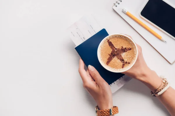Colpo ritagliato di donna che tiene in mano una tazza di caffè con cartello aereo sul tavolo con passaporto, smartphone e biglietto, concetto di viaggio — Foto stock