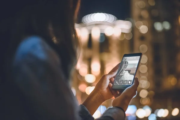 Visão parcial da mulher usando smartphone com bilhetes site na tela luzes da cidade noite no fundo — Fotografia de Stock