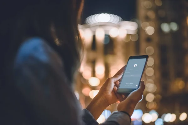 Visão parcial da mulher usando smartphone com logotipo do facebook na tela e noite luzes da cidade no fundo — Fotografia de Stock