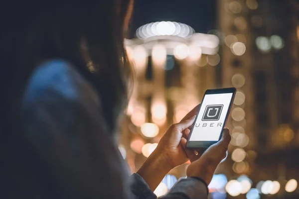Teilansicht der Frau mit Smartphone mit Uber-Logo auf dem Bildschirm und nächtlicher Stadtbeleuchtung im Hintergrund — Stockfoto