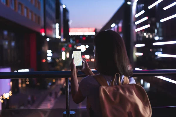 Vista trasera de la mujer con la mochila y el teléfono inteligente con el mapa en la pantalla en las manos de pie en la calle de la ciudad noche - foto de stock