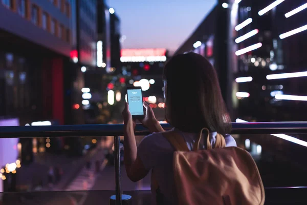 Visão traseira da mulher com mochila e smartphone com logotipo skype na tela em mãos em pé na rua da cidade noite — Fotografia de Stock