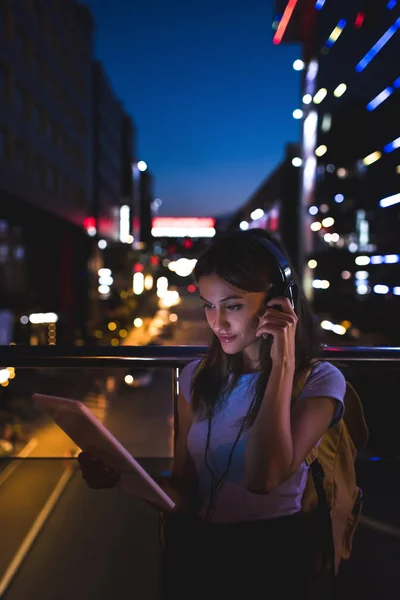 Retrato de mujer en auriculares usando tableta en la calle con luces nocturnas en el fondo - foto de stock