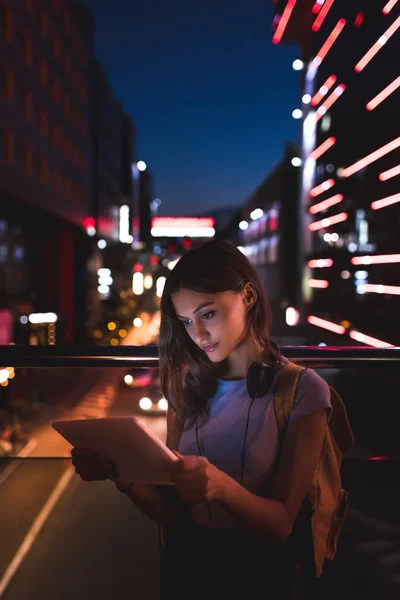 Portrait de jeune femme avec écouteurs utilisant une tablette dans la rue avec des lumières de la ville nocturne sur le fond — Photo de stock