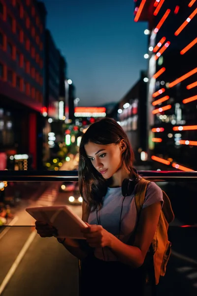 Ritratto di giovane donna con cuffie utilizzando tablet su strada con luci notturne della città su sfondo — Foto stock