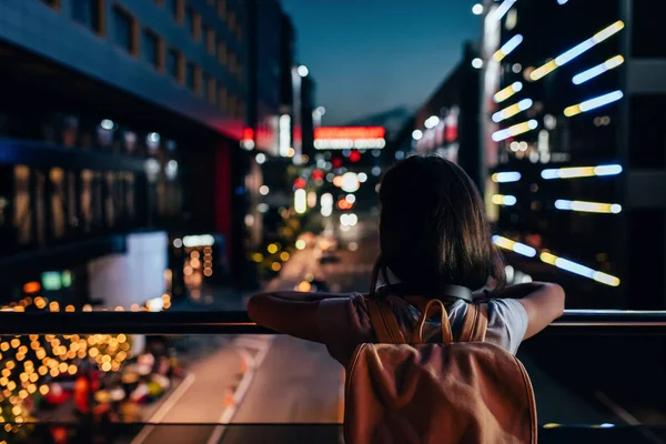 Вид ззаду жінки з рюкзаком, що стоїть на вулиці і дивиться на нічне місто вогні — стокове фото