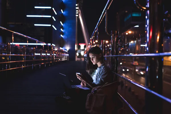 Vista lateral de la mujer joven con el ordenador portátil en las rodillas con el teléfono inteligente con la ciudad de la noche en el fondo - foto de stock