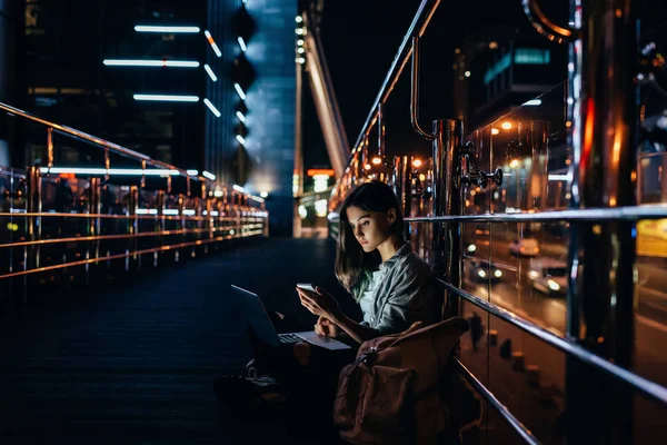 Side view of young woman with laptop on knees using smartphone with night city on background — Stock Photo