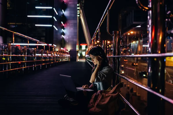 Vista lateral da mulher ouvindo música em fones de ouvido enquanto usa laptop na rua da cidade à noite — Fotografia de Stock