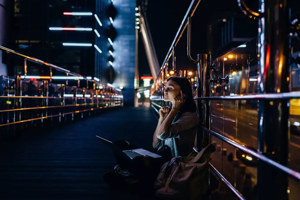 Vista lateral de la mujer escuchando música en auriculares con portátil de rodillas en la calle de la ciudad por la noche - foto de stock