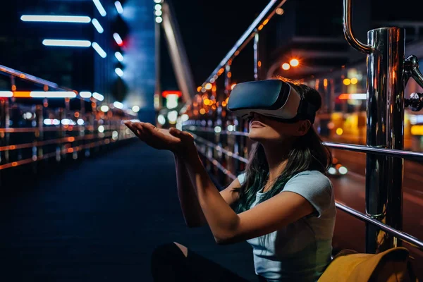 Jovem mulher em realidade virtual fone de ouvido sentado na rua com cidade noturna em segundo plano — Fotografia de Stock