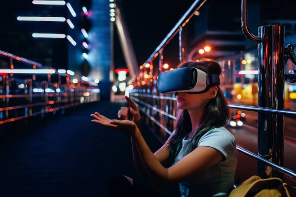 Mujer joven en auriculares de realidad virtual sentado en la calle con la ciudad de la noche en el fondo - foto de stock
