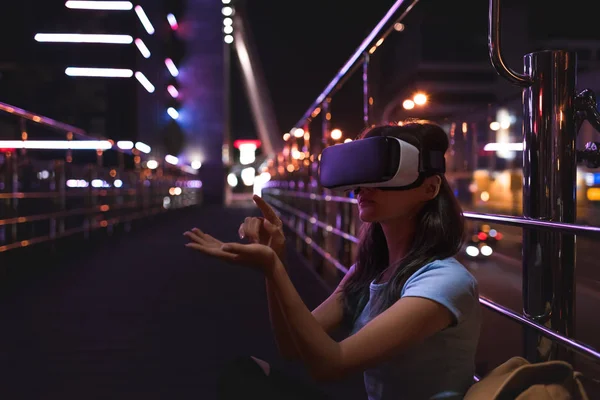 Mujer joven en auriculares de realidad virtual sentado en la calle con la ciudad de la noche en el fondo - foto de stock