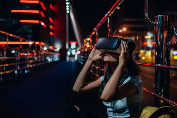 Young woman in virtual reality headset sitting on street with night city on background — Stock Photo