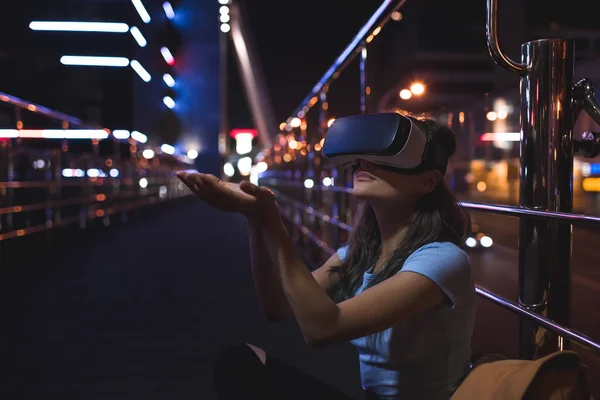 Young woman in virtual reality headset sitting on street with night city on background — Stock Photo