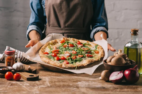 Частичный вид женщины в фартуке, держащей приготовленную пиццу на деревянном столе и ингредиенты — стоковое фото