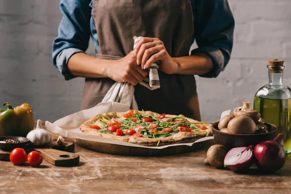 Partial view of woman in apron standing at surface with cooked pizza and ingredients — Stock Photo