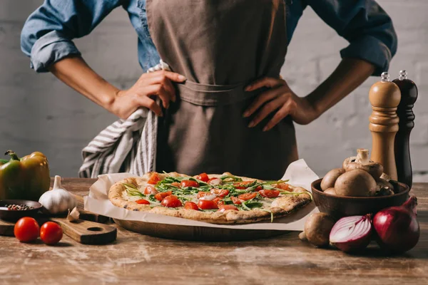 Частичный вид женщины в фартуке, стоящей на поверхности с приготовленной пиццей и ингредиентами — стоковое фото