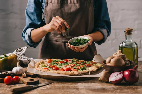 Partial view of woman in apron pouring parsley on cooked pizza at wooden surface — Stock Photo