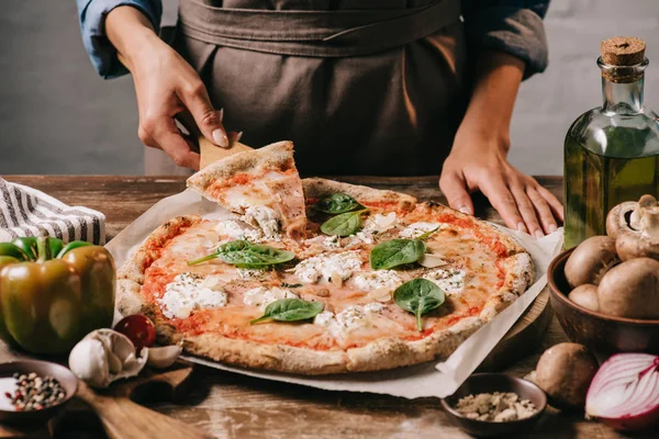 Vue partielle de la femme en tablier prenant un morceau de pizza sur une planche à découper sur une table en bois — Photo de stock