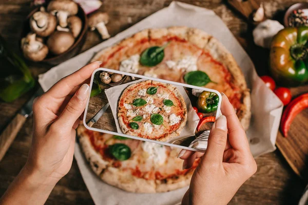 Foto recortada de bloguero de alimentos tomando fotos de pizza cocida en papel de hornear en la superficie de madera - foto de stock