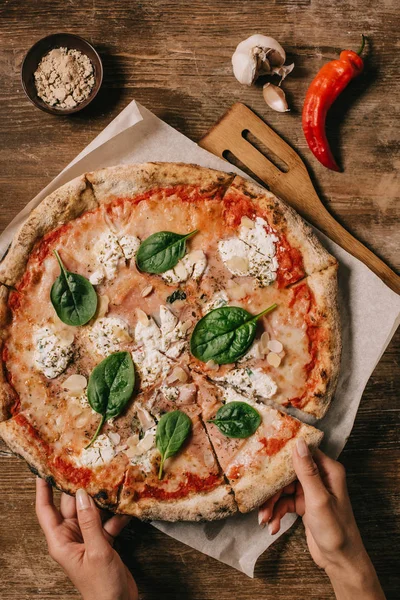 Teilansicht einer Frau, die ein Stück Pizza auf einem Schneidebrett auf einer hölzernen Tischplatte nimmt — Stockfoto
