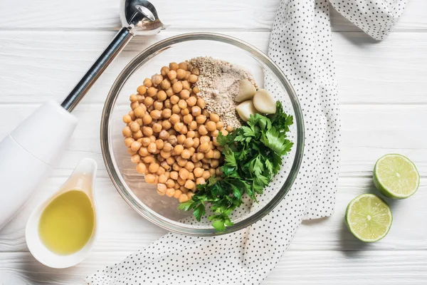 Flat lay with chickpeas, parsley and spices ingredients for hummus in bowl, blender and lime pieces on wooden surface — Stock Photo