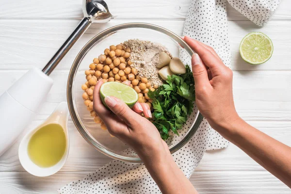 Cropped shot of woman, ingredients for hummus in bowl, olive oil and blender on wooden tabletop — Stock Photo