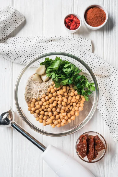 Acostado plano con garbanzos y otros ingredientes para hummus en un tazón, licuadora y tomates secos sobre una mesa de madera - foto de stock