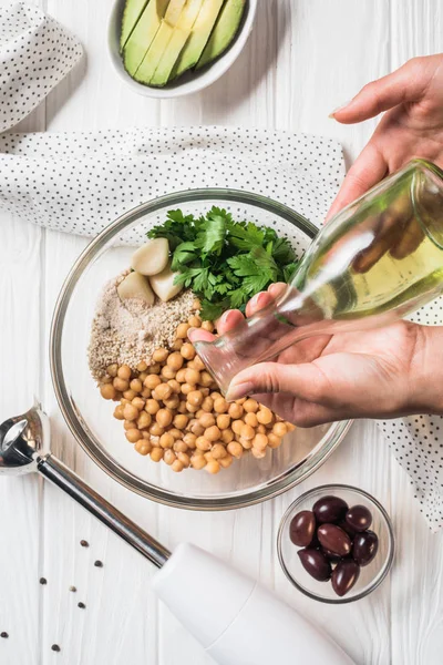 Colpo ritagliato di donna versando olio d'oliva in ciotola con ceci e altri ingredienti per hummus su un tavolo di legno — Foto stock