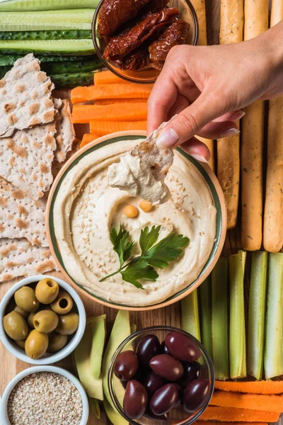 Partial view of woman and arranged hummus in bowl, pita bread, cut vegetables, dried tomatoes and olives — Stock Photo
