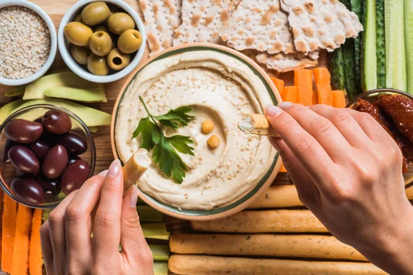 Partial view of woman and arranged hummus in bowl, pita bread, cut vegetables, dried tomatoes and olives — Stock Photo