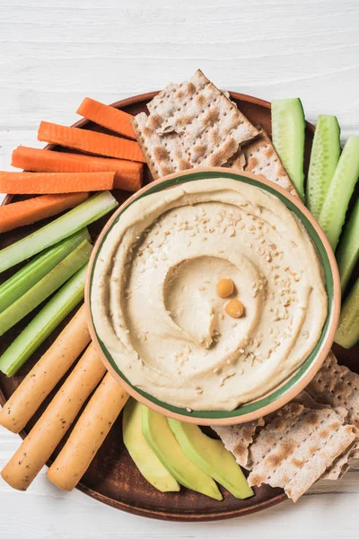 Vue de dessus du houmous dans un bol avec des tranches de légumes coupés disposés, des bâtonnets de pain et du pain pita sur la surface en bois — Photo de stock