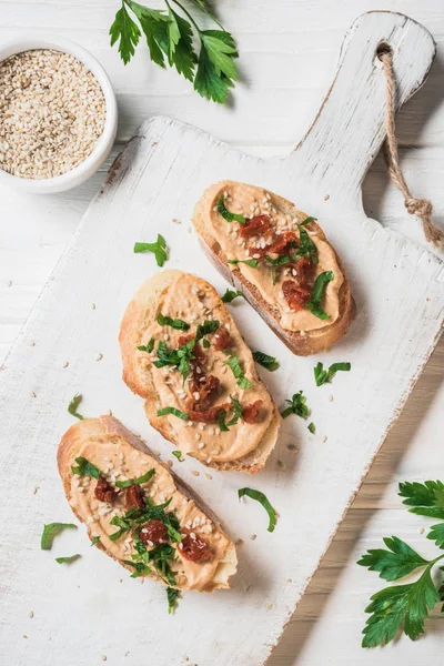 Top view of pieces of bread with hummus and dried tomatoes on cutting board on wooden tabletop — Stock Photo