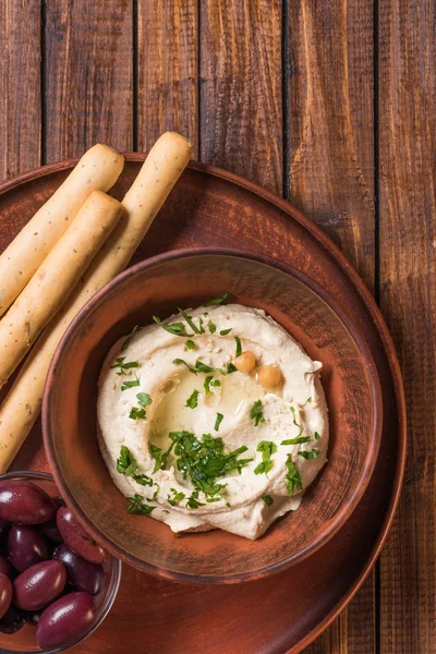 Vista superior de aceitunas, palitos de pan y hummus con perejil y garbanzos en tazón sobre superficie de madera - foto de stock