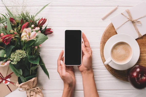 Foto recortada de la mujer sosteniendo teléfono inteligente con pantalla en blanco en la superficie con ramo envuelto de flores y taza de café - foto de stock